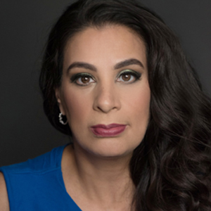 Headshot of comedian Maysoon Zayid, a brunette woman with long hair, gold earrings and red lips. She is wearing a sleeveless blue top.